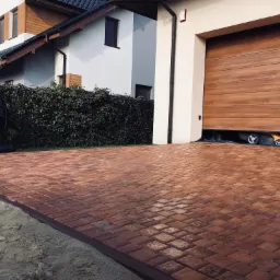 Ułożona, czerwono-brązowa kostka brukowa na podjeździe przed garażem, widoczny piasek przy krawędzi, w tle dom z zielonym żywopłotem i częściowo otwarta brama garażowa z widocznymi kołami samochodu.