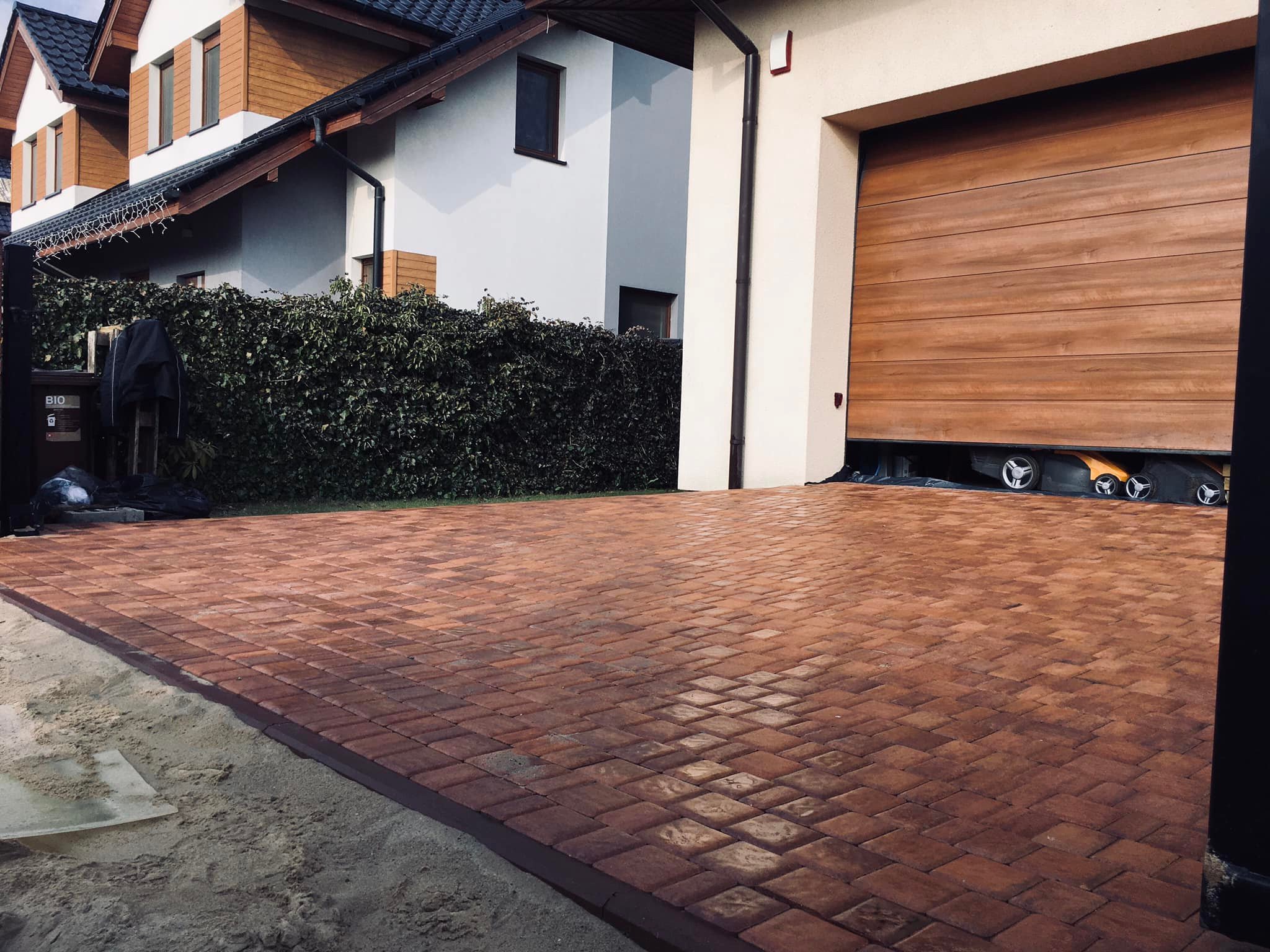 Ułożona, czerwono-brązowa kostka brukowa na podjeździe przed garażem, widoczny piasek przy krawędzi, w tle dom z zielonym żywopłotem i częściowo otwarta brama garażowa z widocznymi kołami samochodu.