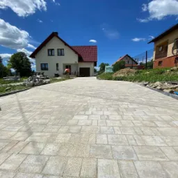 Szeroki podjazd z jasnoszarej kostki brukowej ułożonej we wzór prostokątny, widok z perspektywy chodnika, w tle dom jednorodzinny z czerwoną dachówką i betoniarka.