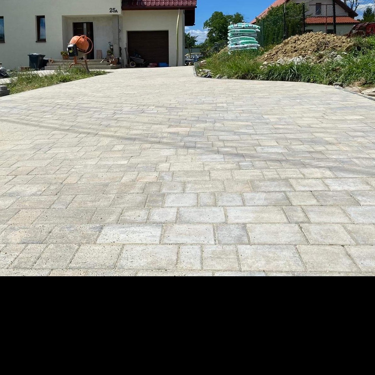 Szeroki podjazd wyłożony jasnoszarą kostką brukową w regularny wzór, z widocznym mieszalnikiem do betonu i stertą worków w tle.