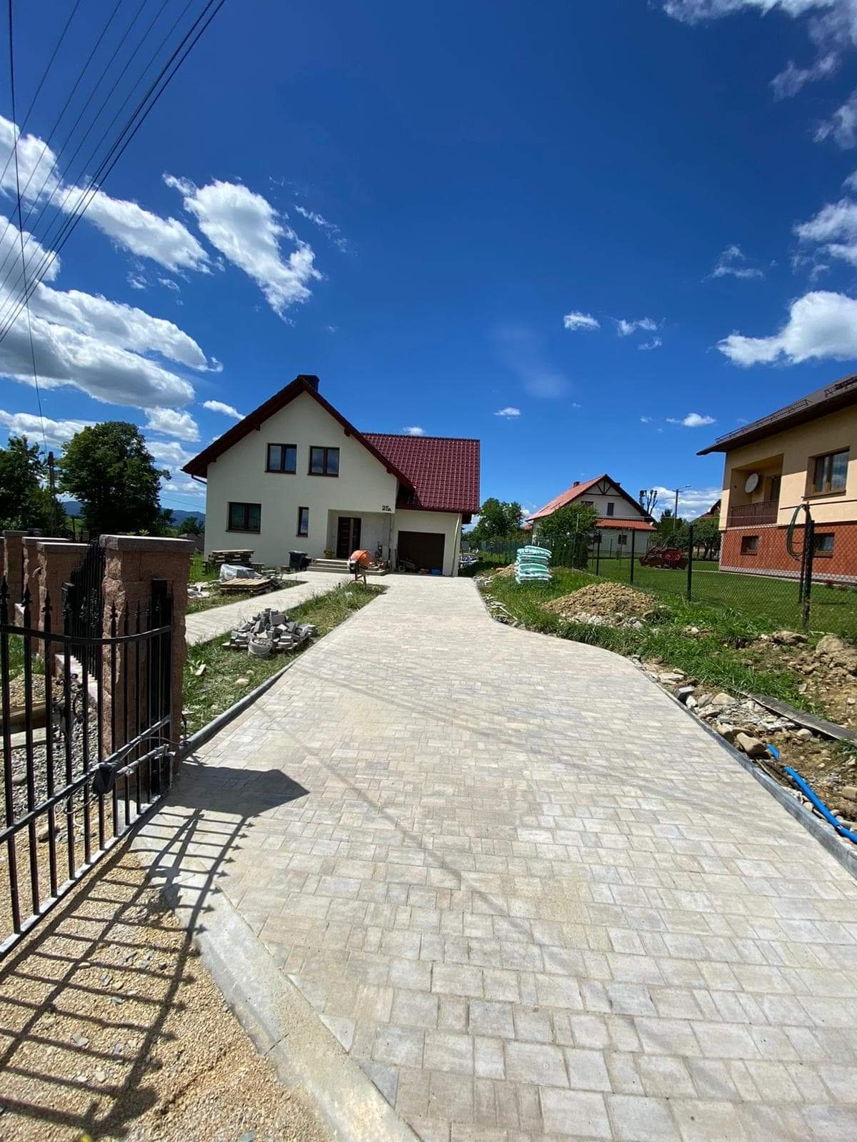 Nowa, jasna nawierzchnia z kostki brukowej prowadząca do domu z czerwonym dachem, widok zza czarnej, metalowej bramy, błękitne niebo z białymi obłokami w tle.