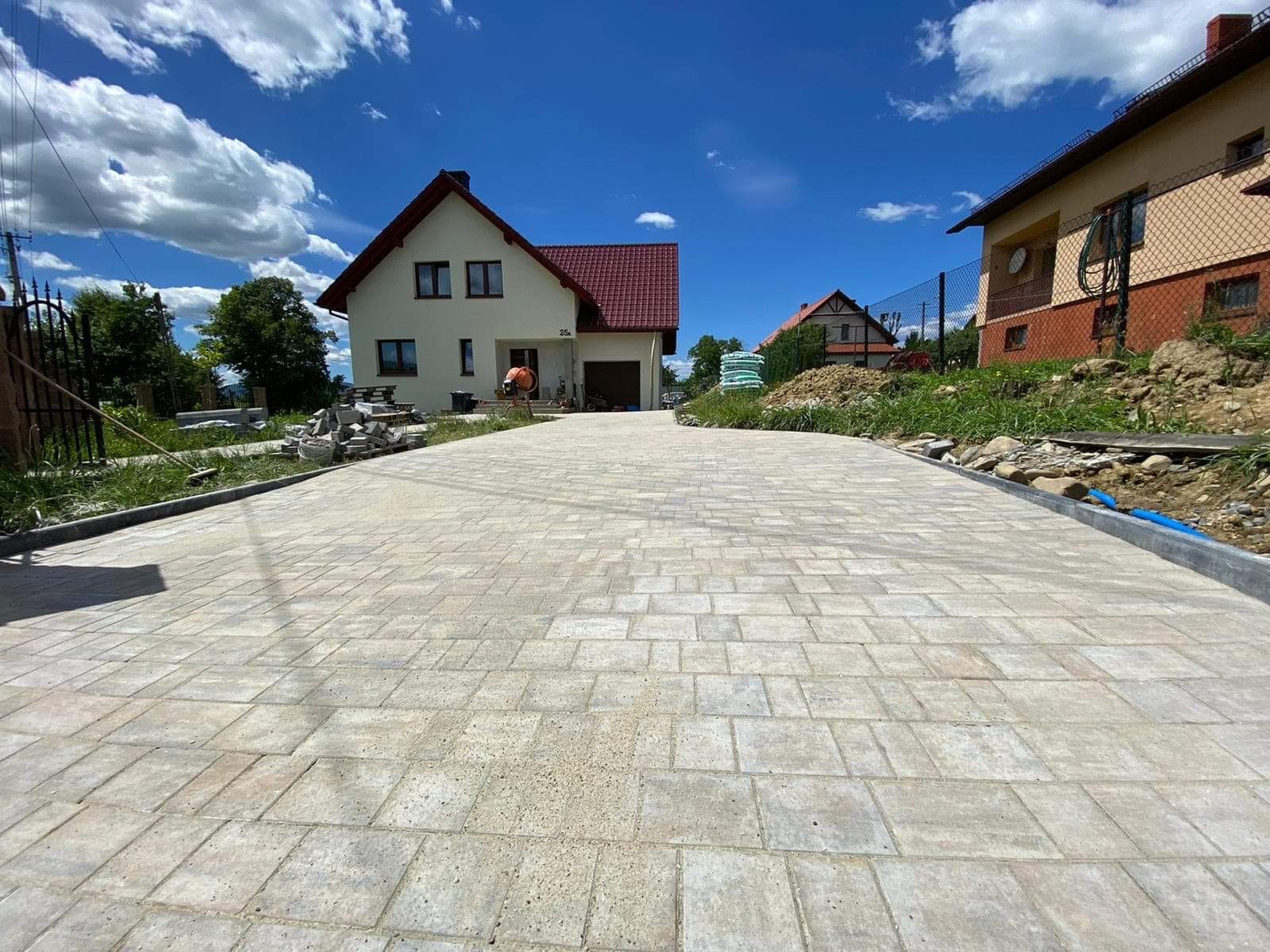 Szeroki podjazd z jasnoszarej kostki brukowej ułożonej we wzór prostokątny, widok z perspektywy chodnika, w tle dom jednorodzinny z czerwoną dachówką i betoniarka.
