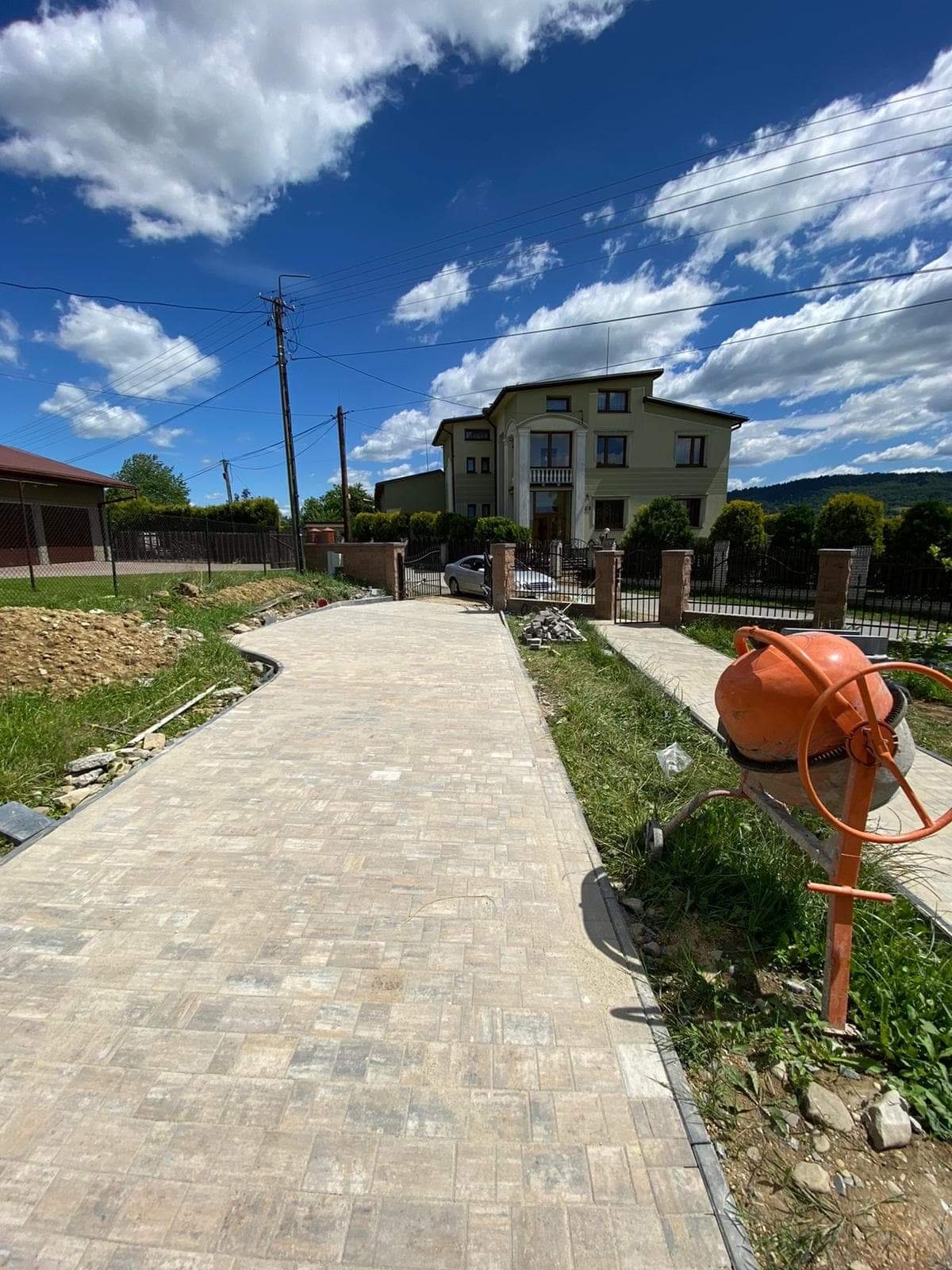 Nowo ułożony, jasnoszary chodnik z kostki brukowej prowadzący do domu z widoczną betoniarką i przygotowanym terenem pod dalsze prace.