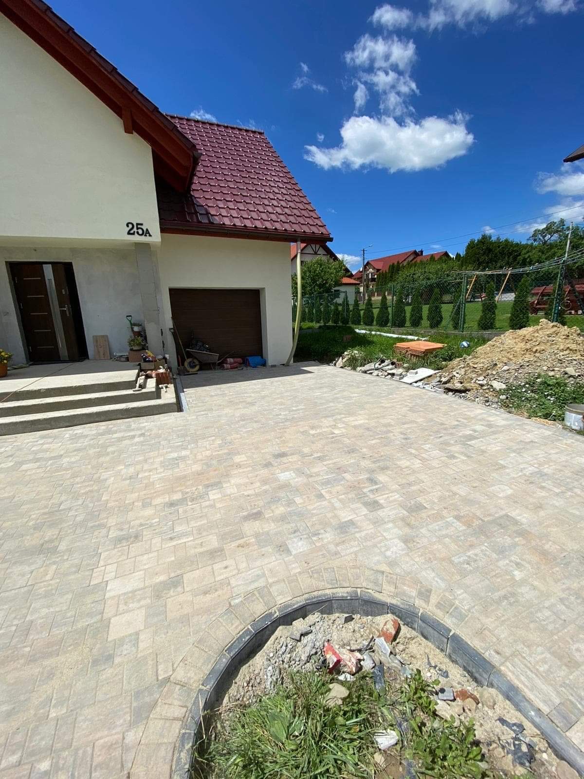 Ułożona kostka brukowa na podjeździe domu z widocznym numerem 25A, z niezakończonym obrzeżem i stertą ziemi w tle.