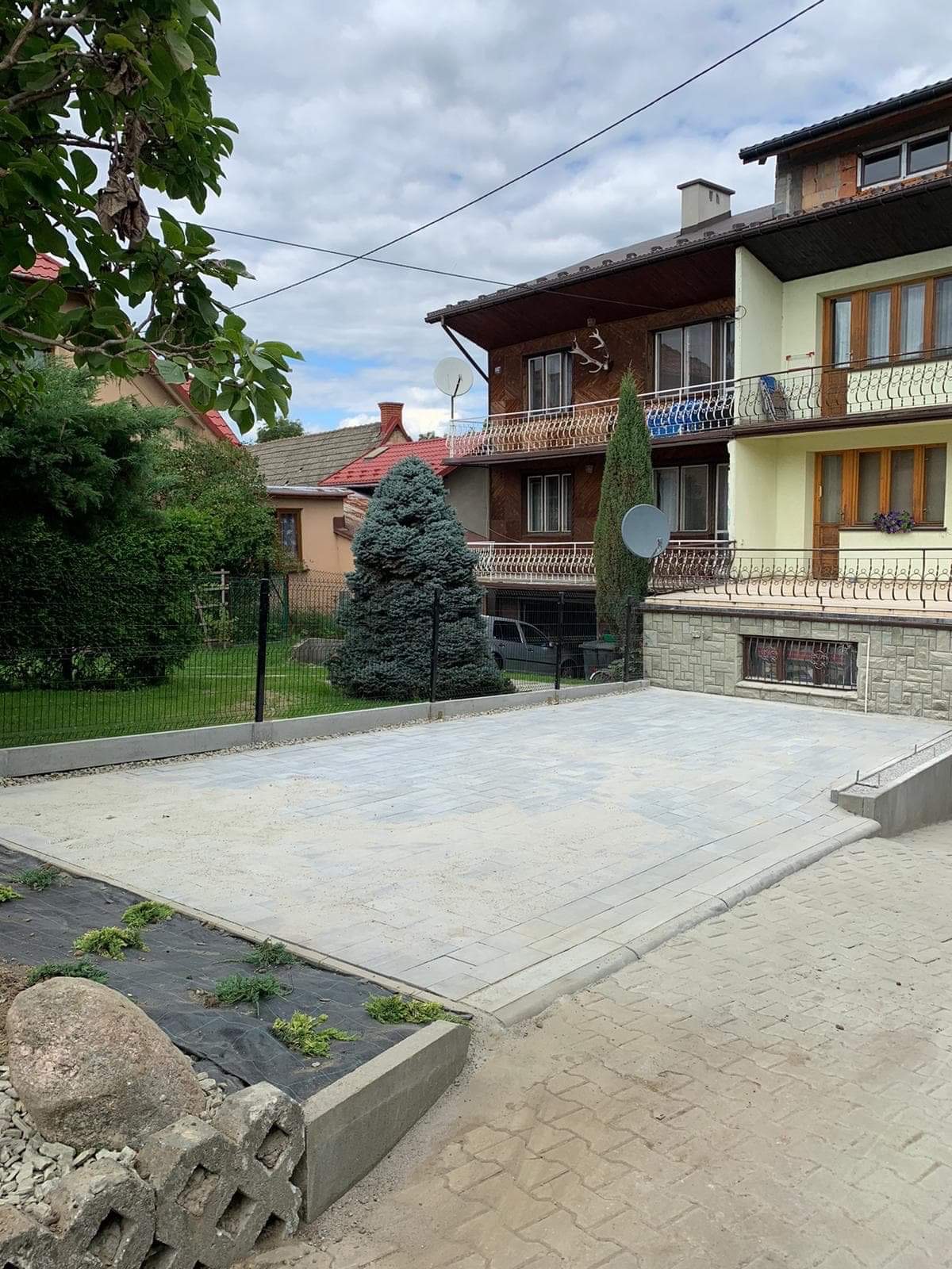 Nowo ułożony, szary podjazd z kostki brukowej przed domem jednorodzinnym, widoczne obrzeża, trawnik z młodymi roślinami i kamieniami, w tle budynki mieszkalne.