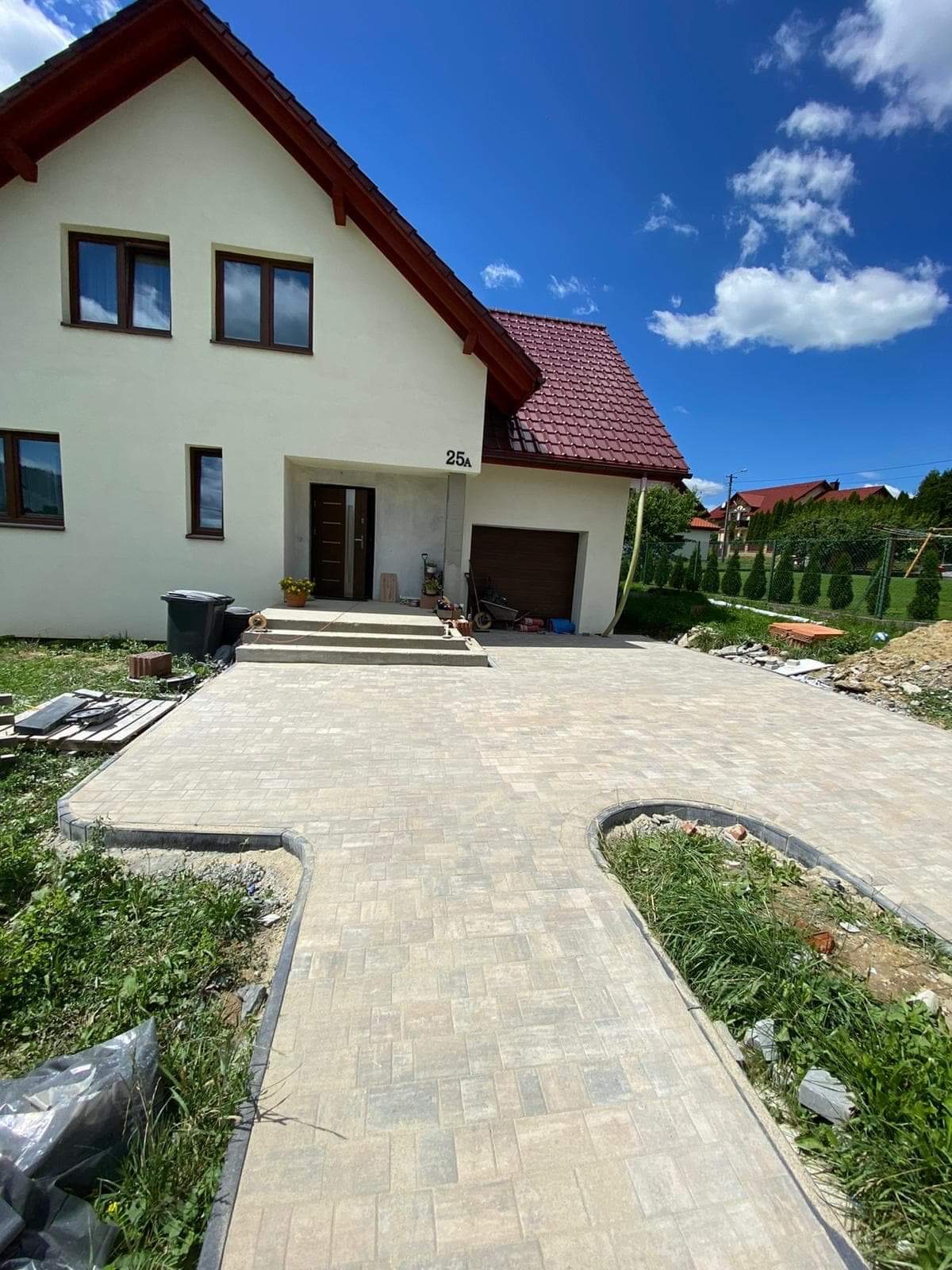 Nowy podjazd z kostki brukowej przed domem jednorodzinnym z czerwonym dachem, widok na numer domu 25A i częściowo otwarty garaż.