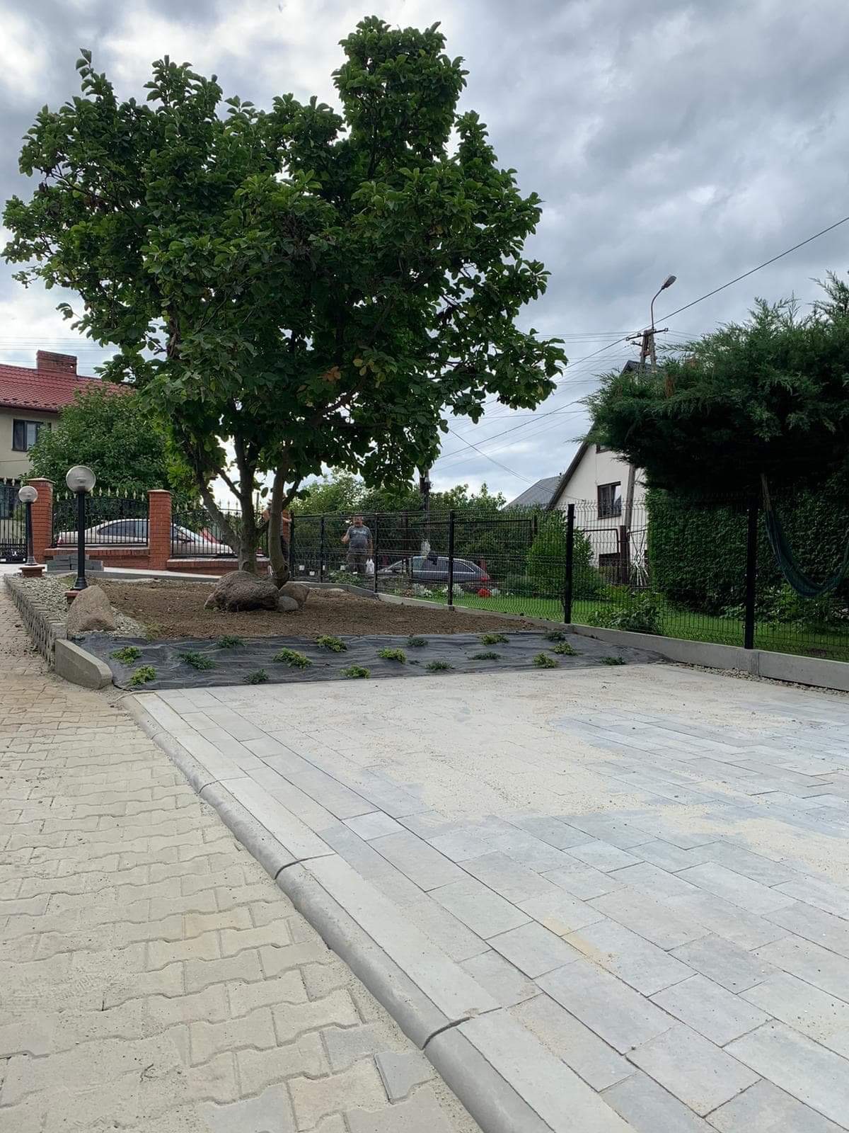 Świeżo ułożona, jasnoszara kostka brukowa na podjeździe, z obrzeżami i przygotowaną rabatą z agrowłókniną pod nasadzenia, w tle ogrodzenie z siatki.