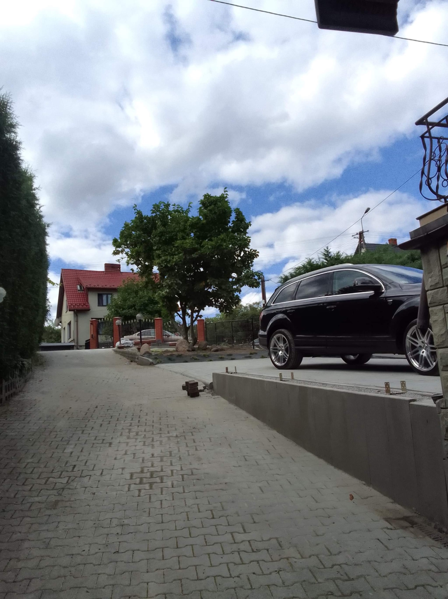 Ułożona szara kostka brukowa na podjeździe z widokiem na dom z czerwoną dachówką i czarne auto zaparkowane obok nowego betonowego murku.