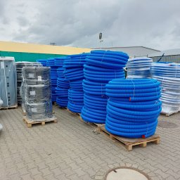 Apis Sp. z O.O. Grupa Sbs - Magazynowane zwoje niebieskich rur karbowanych na paletach, obok zbiorniki i inne elementy instalacji, na zewnątrz pod pochmurnym niebem.