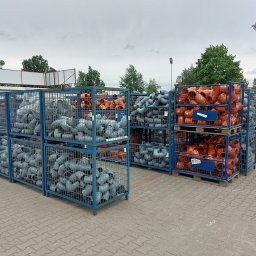 Apis Sp. z O.O. Grupa Sbs - Magazyn rur kanalizacyjnych PCV w różnych kolorach, ułożonych w metalowych koszach na paletach na zewnątrz.