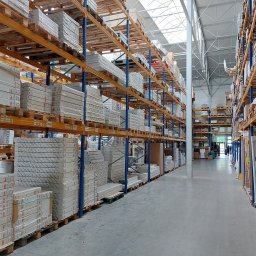 Apis Sp. z O.O. Grupa Sbs - Magazyn z regałami wypełnionymi zapakowanymi urządzeniami sanitarnymi i grzewczymi, widoczne marki KOŁO, OMRO i inne, oświetlenie górne.