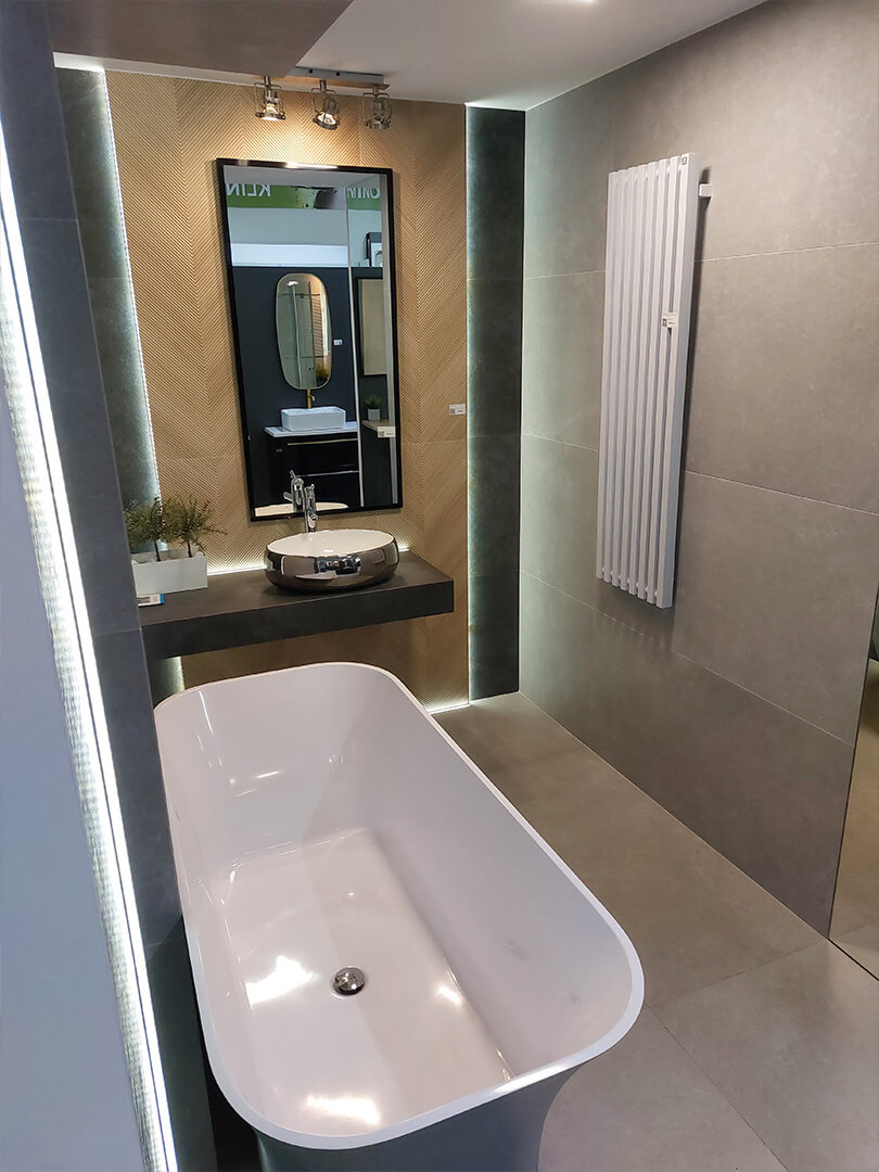 Nowoczesna łazienka z wolnostojącą wanną, umywalką nablatową i grzejnikiem drabinkowym na tle szarych płytek. Widoczne oświetlenie LED wzdłuż krawędzi ścian i lustra.