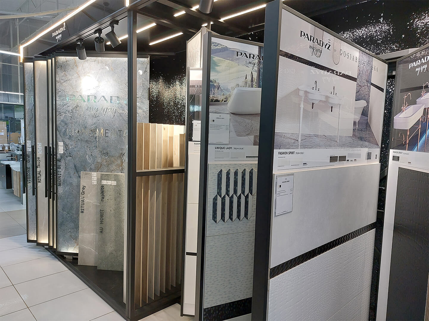 Ekspozycja różnych wzorów i kolorów płytek ceramicznych marki Paradyż w salonie, prezentowanych na metalowym stojaku z oświetleniem LED, zawierająca aranżacje łazienek z umywalkami i wannami.
