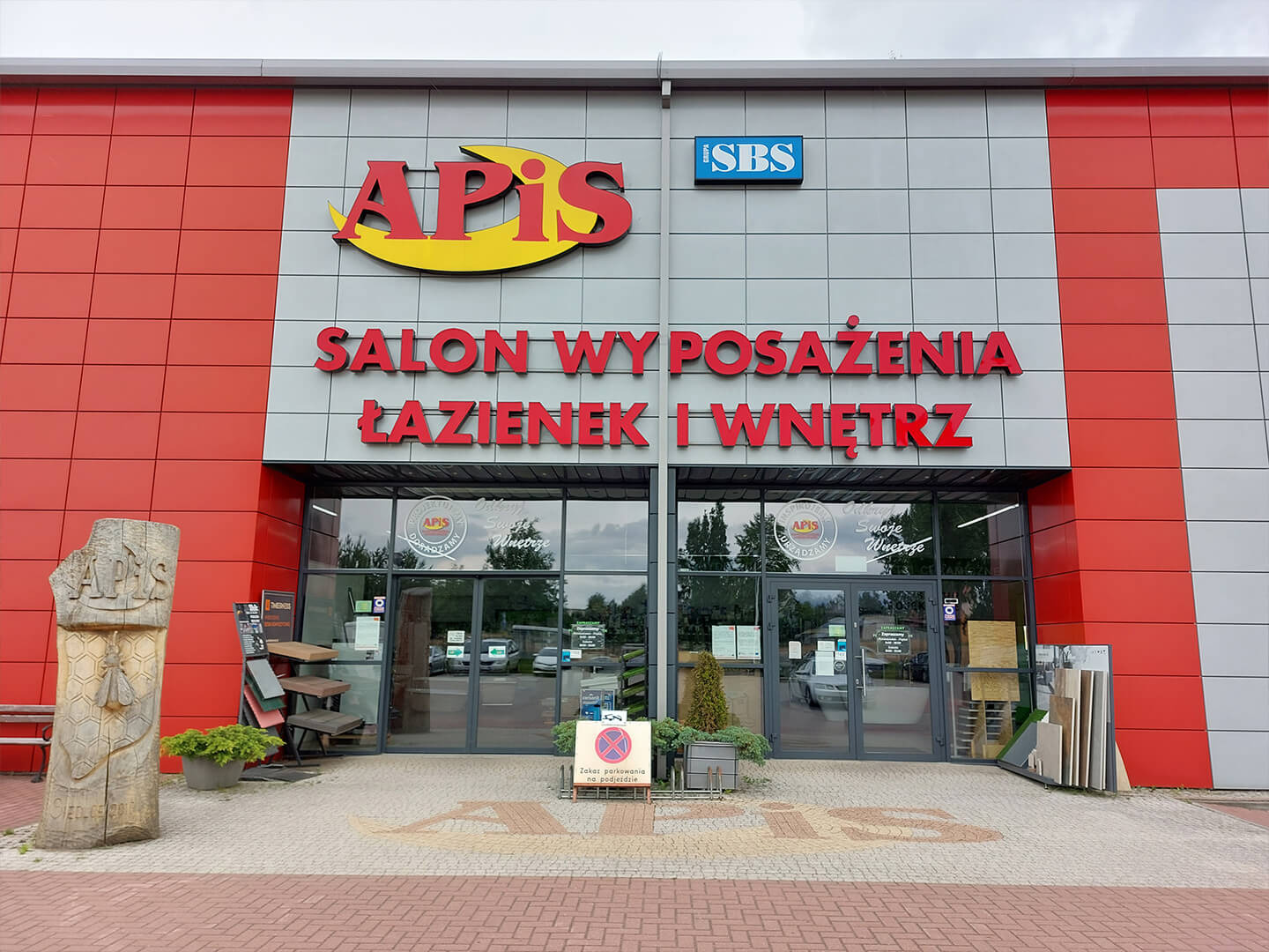 Fasada budynku z napisem 'APIS SALON WYPOSAŻENIA ŁAZIENEK I WNĘTRZ' nad wejściem, z dekoracyjną rzeźbą drewnianą z motywem pszczoły po lewej stronie.
