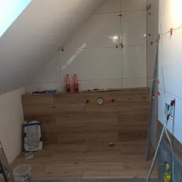 Łazienka na poddaszu w trakcie remontu: dolna część ściany wyłożona panelami imitującymi drewno, g&oacute;rna część w trakcie układania jasnych płytek, widoczne krzyżyki dystansowe i poziomica.