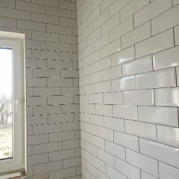 Ściana wyłożona białymi, prostokątnymi płytkami z ciemną fugą, widoczne krzyżyki dystansowe między płytkami, surowy betonowy sufit i okno z widokiem na drzewa.