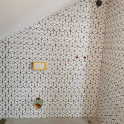 Ściana łazienki w Koszalinie wyłożona biało-czarnymi płytkami w geometryczny wzór 3D kostki, widoczne otwory na instalacje, żółta instrukcja na ścianie i surowa podłoga.