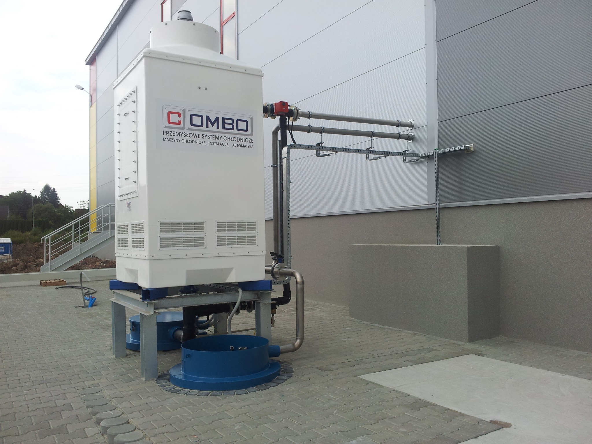 Biała przemysłowa chłodnica COMBO z rurami na zewnątrz budynku z szarej blachy falistej, widoczne elementy instalacji i logo firmy.
