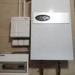 Biały, wiszący kocioł gazowy z elektronicznym panelem sterowania, widoczne zawory odcinające, obok skrzynka elektryczna i peszle instalacyjne na beżowej ścianie.