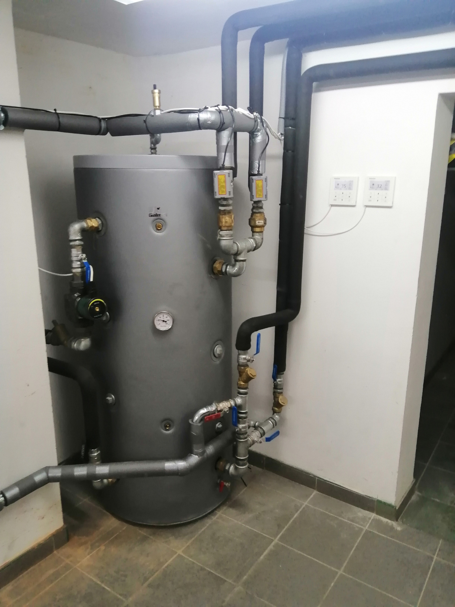 Szary zasobnik ciepłej wody użytkowej z podłączonymi rurami i zaworami w kotłowni, widoczne termostaty na ścianie, rury zaizolowane termicznie.