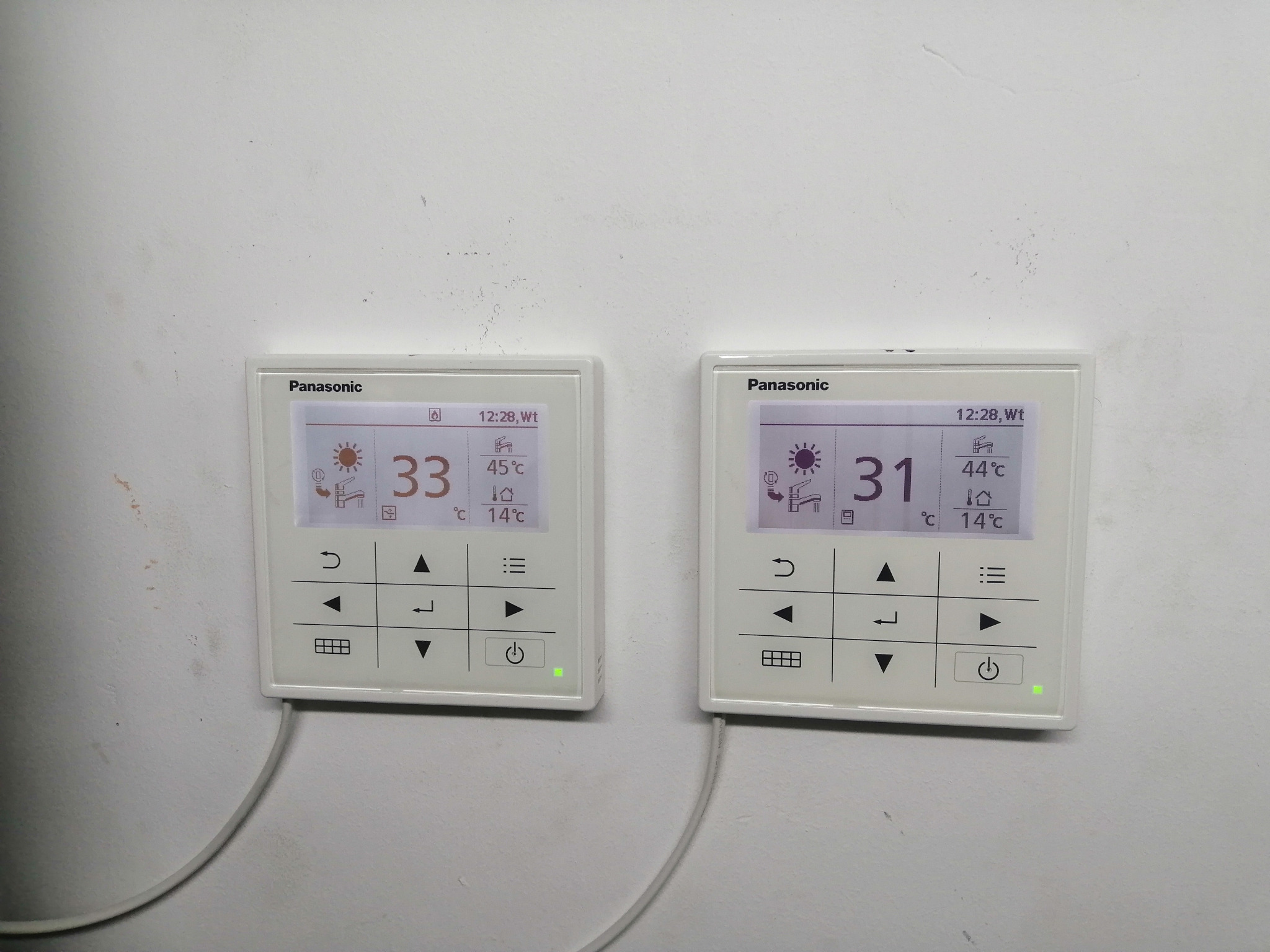 Dwa białe, prostokątne panele sterowania Panasonic zamontowane na pomalowanej ścianie. Wyświetlacze pokazują temperaturę 33°C i 31°C, datę 12:28 oraz ikonę słońca. Zielone diody LED świecą się...