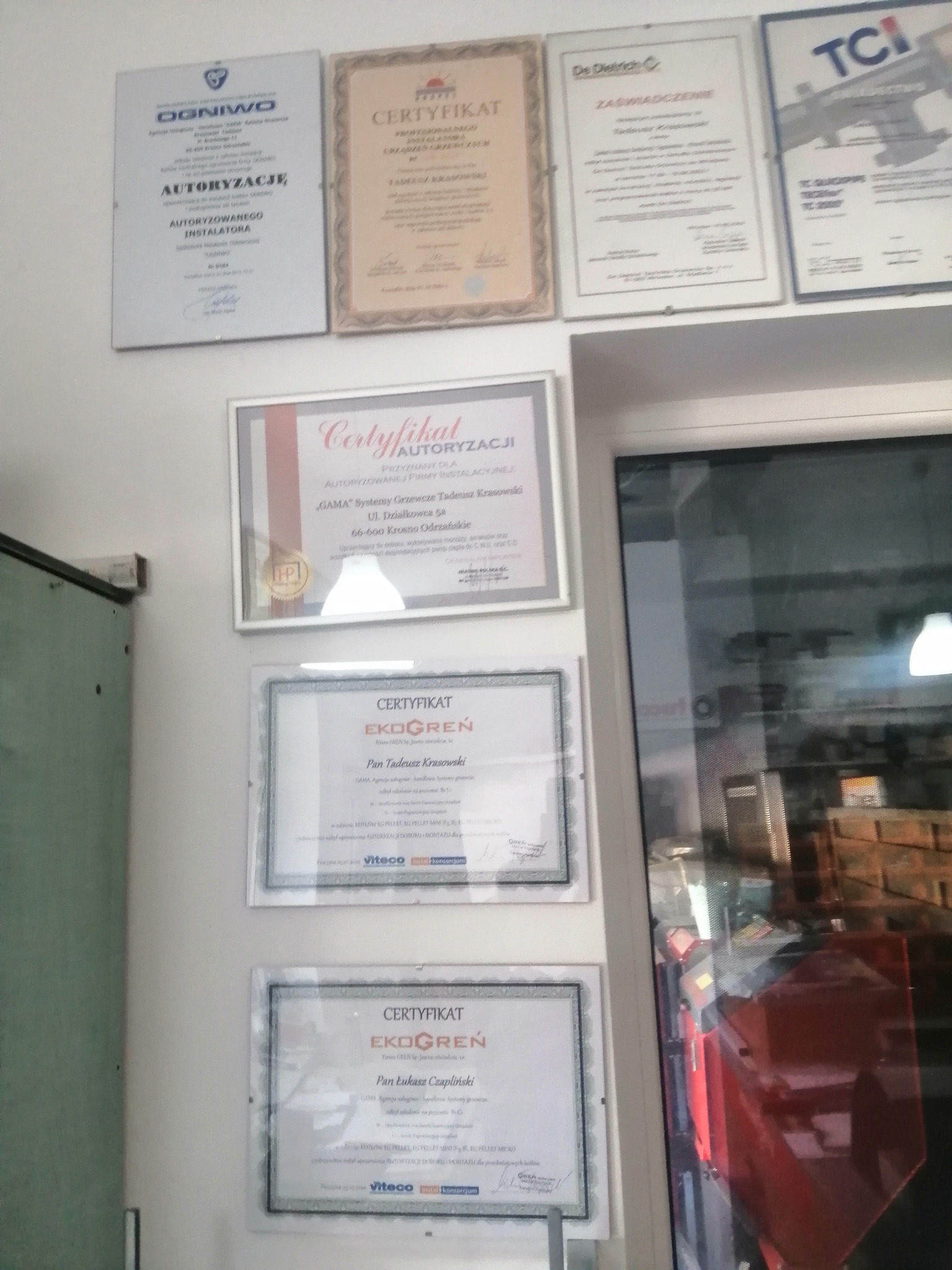 Ściana z powieszonymi certyfikatami i autoryzacjami firm z branży instalacyjnej, w tym certyfikaty 'Ogniwo', 'De Dietrich', 'TCI', 'Certyfikat Autoryzacji', 'Ekogreń', z widocznymi nazwami firm...