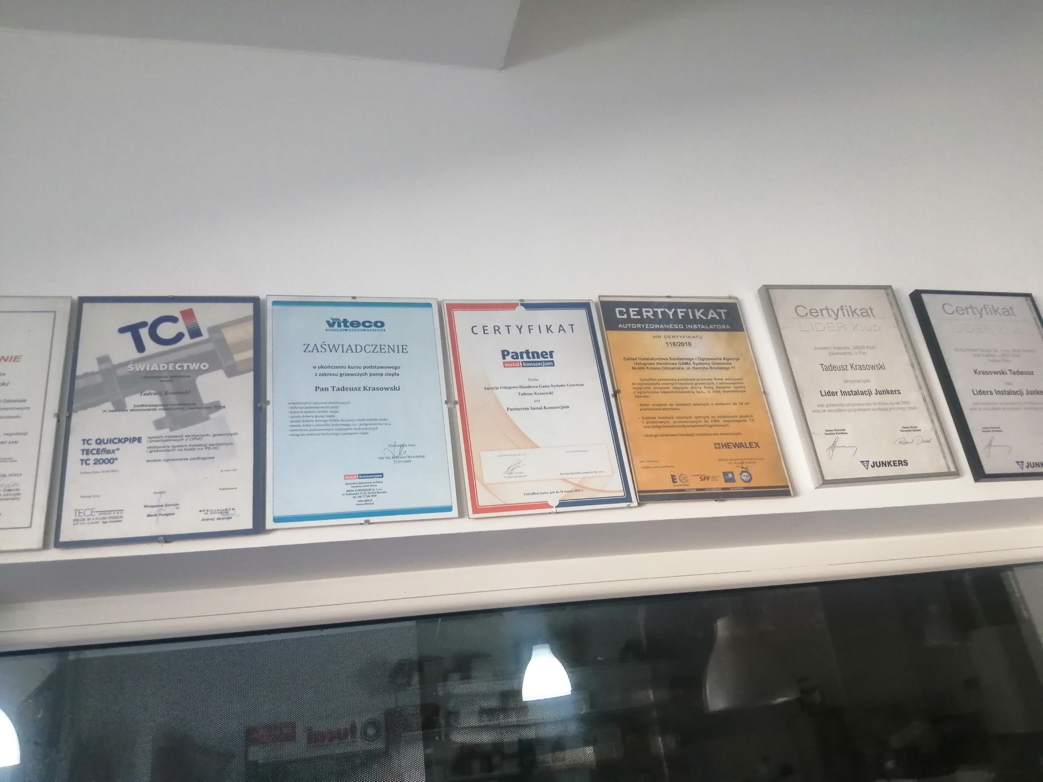 Ściana z certyfikatami i świadectwami firmy instalacyjnej, widoczne ramy z nazwami marek i nazwiskami.