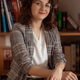 Promed Psychoterapia - Portret uśmiechniętej kobiety w okularach i marynarce w kratę, siedzącej na tle regału z książkami. Ciepłe oświetlenie, profesjonalny wygląd.