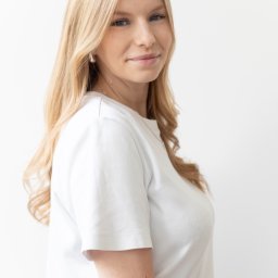 Promed Psychoterapia - Portret kobiety z długimi blond włosami, ubrana w białą koszulkę, spoglądającej w bok na jasnym tle. Widoczny delikatny makijaż i subtelna biżuteria.