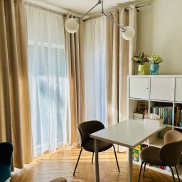 Promed Psychoterapia - Jasny gabinet psychoterapeuty: białe biurko, krzesła, regał z książkami, okno z zasłonami. Dwie rośliny w doniczkach w kształcie głowy. Przytulne i profesjonalne wnętrze.