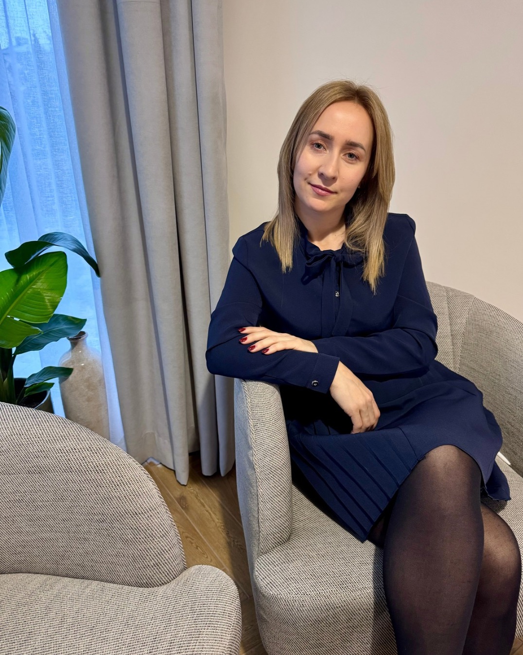 Kobieta w granatowej sukience siedzi w fotelu na tle okna z zasłonami i rośliną doniczkową. Uśmiech, kontakt wzrokowy, profesjonalny wygląd.