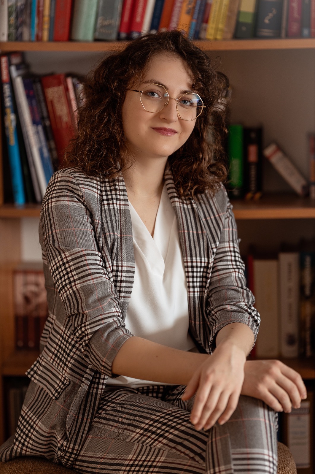 Portret uśmiechniętej kobiety w okularach i marynarce w kratę, siedzącej na tle regału z książkami. Ciepłe oświetlenie, profesjonalny wygląd.