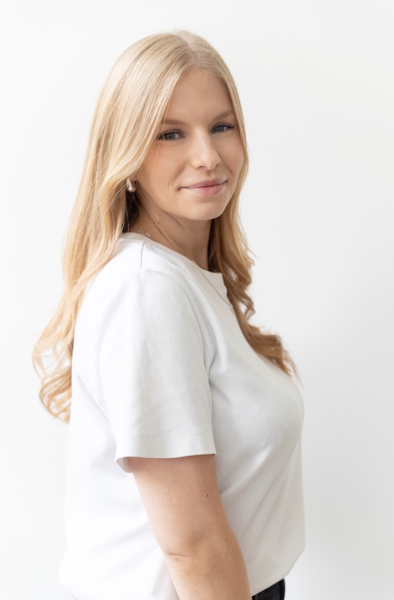 Portret kobiety z długimi blond włosami, ubrana w białą koszulkę, spoglądającej w bok na jasnym tle. Widoczny delikatny makijaż i subtelna biżuteria.