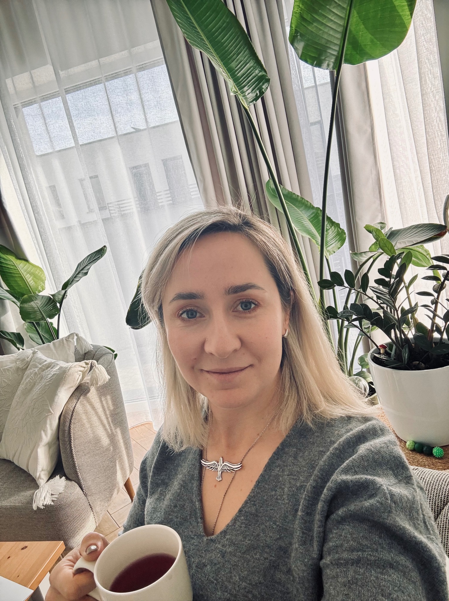 Kobieta z blond włosami pije herbatę w jasnym pokoju z roślinami doniczkowymi i dużym oknem. Uśmiechnięta twarz, srebrny naszyjnik z motywem ptaka.