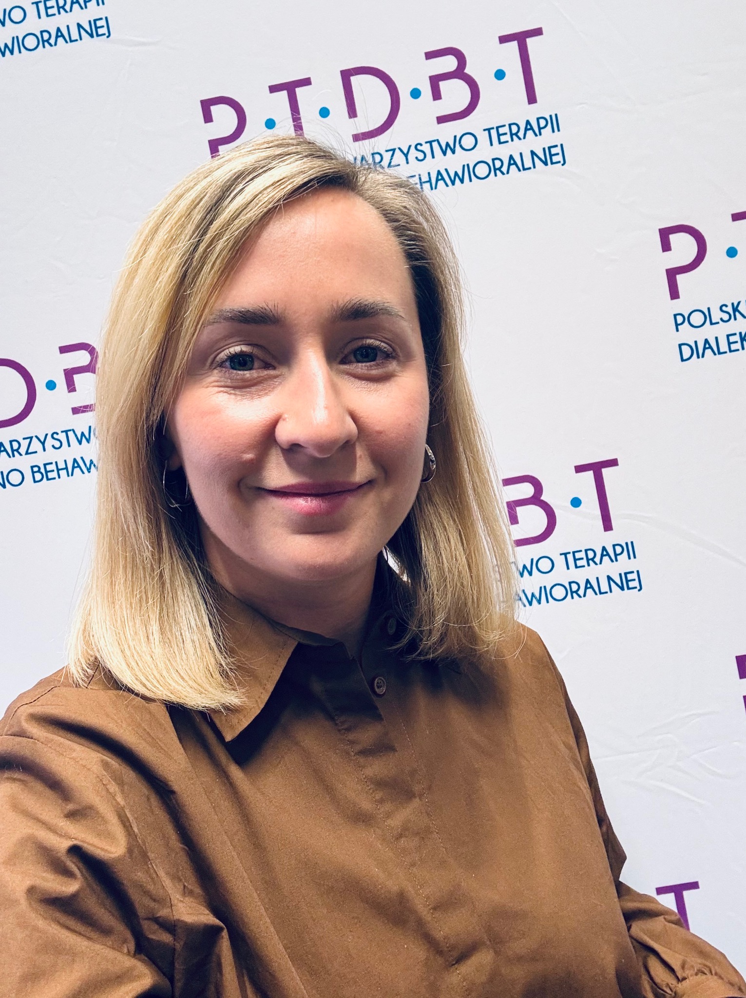 Portret uśmiechniętej blondynki w brązowej koszuli na tle logo P.T.D.B.T. (Polskie Towarzystwo Terapii Dialektyczno-Behawioralnej).