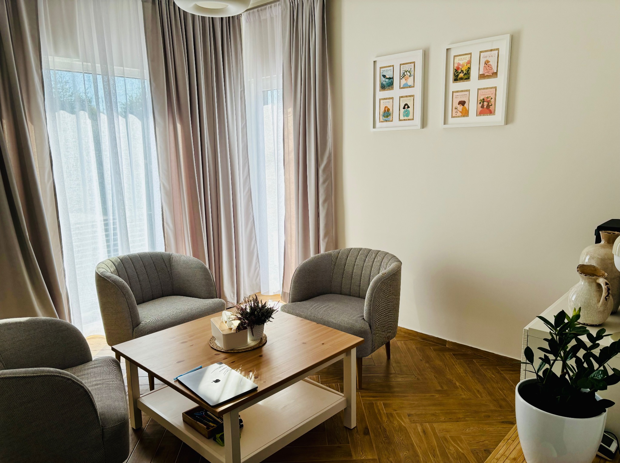 Jasny pokój z trzema fotelami, drewnianym stolikiem kawowym z laptopem i kwiatami. Na ścianie wiszą dwie oprawione grafiki. Okno z zasłonami w tle.