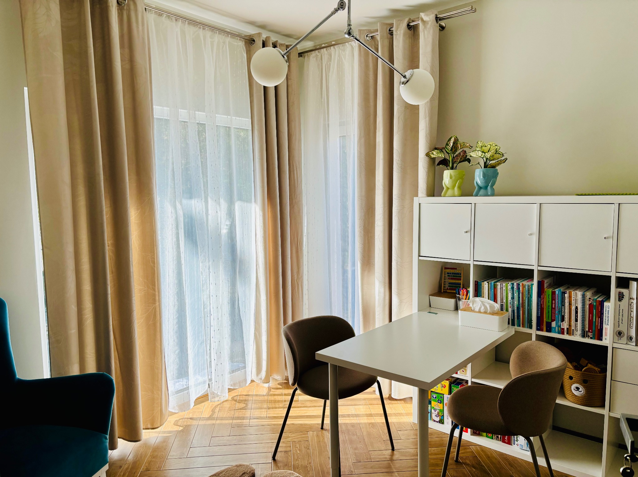 Jasny gabinet psychoterapeuty: białe biurko, krzesła, regał z książkami, okno z zasłonami. Dwie rośliny w doniczkach w kształcie głowy. Przytulne i profesjonalne wnętrze.