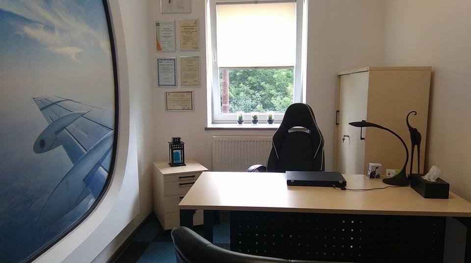 Gabinet psychoterapeuty z biurkiem, krzesłem gamingowym, ścianą z certyfikatami i okrągłym obrazem skrzydła samolotu na ścianie.