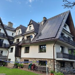 OZEPODHALE Dawid Knutelski, Zbigniew Knutelski spółka cywilna - Elewacja budynku z panelami fotowoltaicznymi na dachu, widok z ogrodu z siatką do gry w siatkówkę i osobą w tle. Biała fasada i kamienne wykończenia.