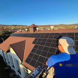 OZEPODHALE Dawid Knutelski, Zbigniew Knutelski spółka cywilna - Brązowy dach budynku z panelami słonecznymi w słoneczny dzień, widok z góry z pracownikiem w niebieskiej koszulce z logo firmy.