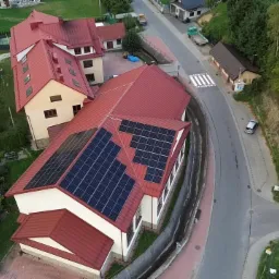 Widok z g&oacute;ry na budynek z czerwonym dachem, na kt&oacute;rym zamontowane są panele słoneczne. W tle zabudowa wiejska i droga.