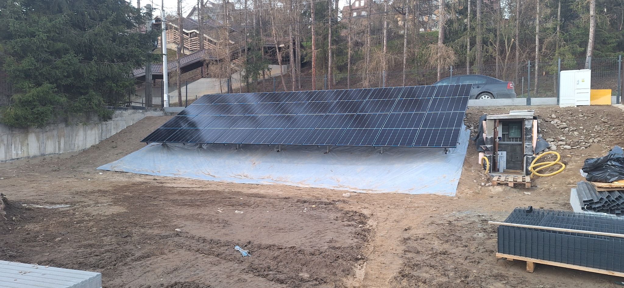 Instalacja paneli słonecznych na gruncie, widoczny rozdzielacz i elementy montażowe. W tle drzewa i domy. Ujęcie z poziomu gruntu.