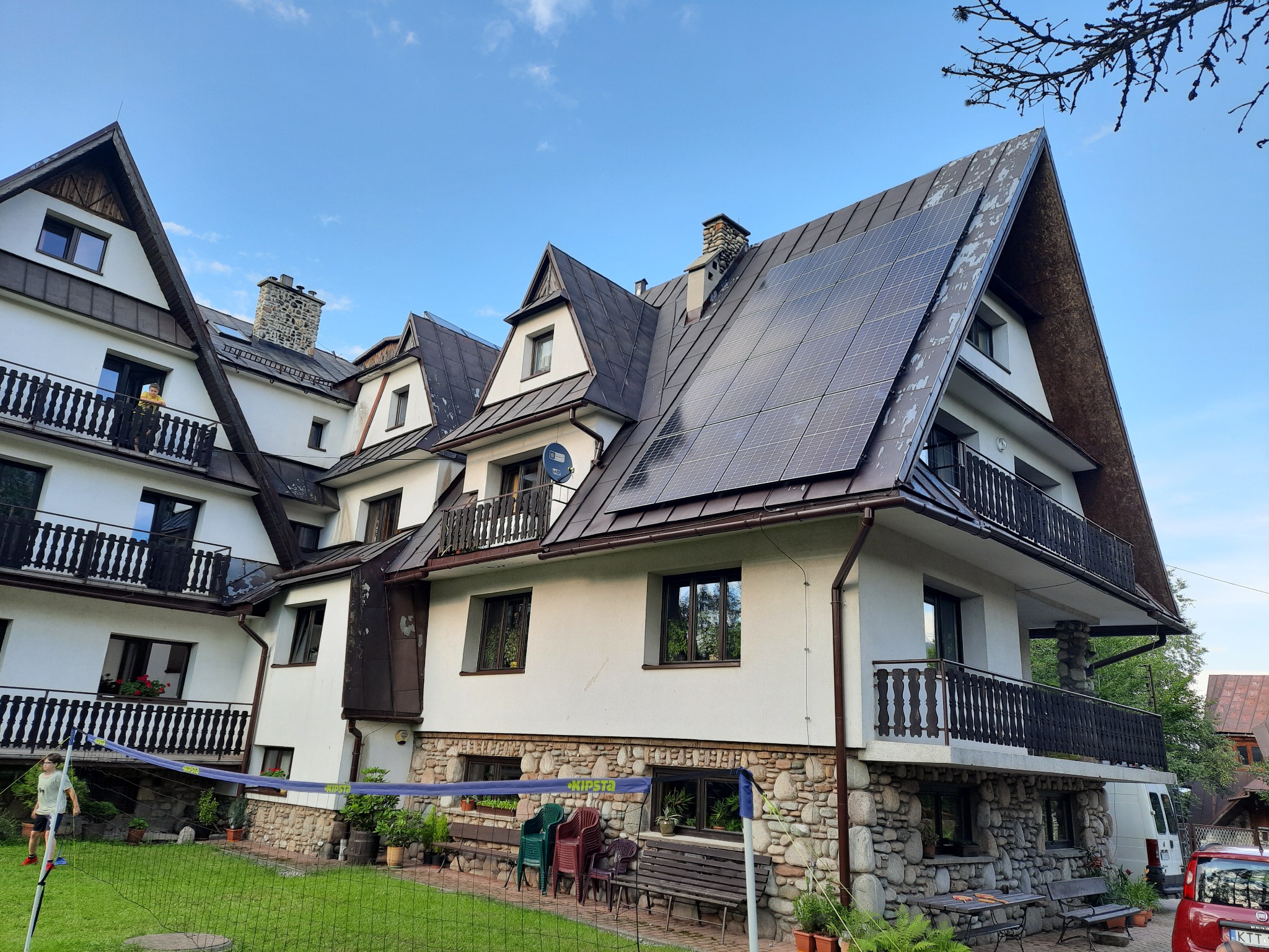 Elewacja budynku z panelami fotowoltaicznymi na dachu, widok z ogrodu z siatką do gry w siatkówkę i osobą w tle. Biała fasada i kamienne wykończenia.