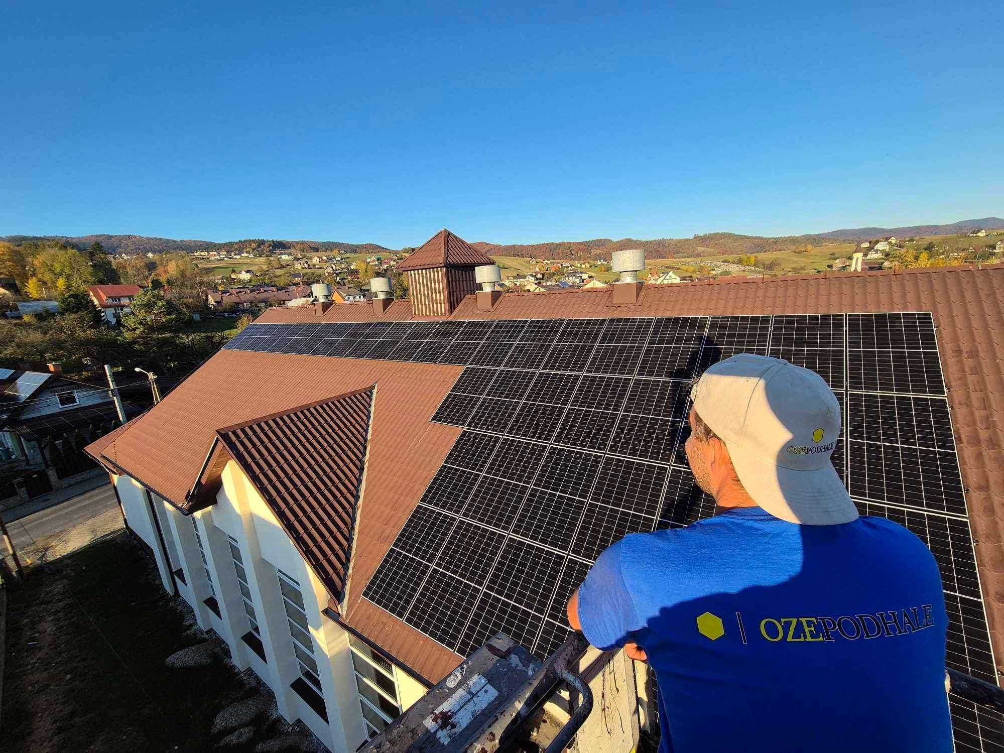 Brązowy dach budynku z panelami słonecznymi w słoneczny dzień, widok z góry z pracownikiem w niebieskiej koszulce z logo firmy.