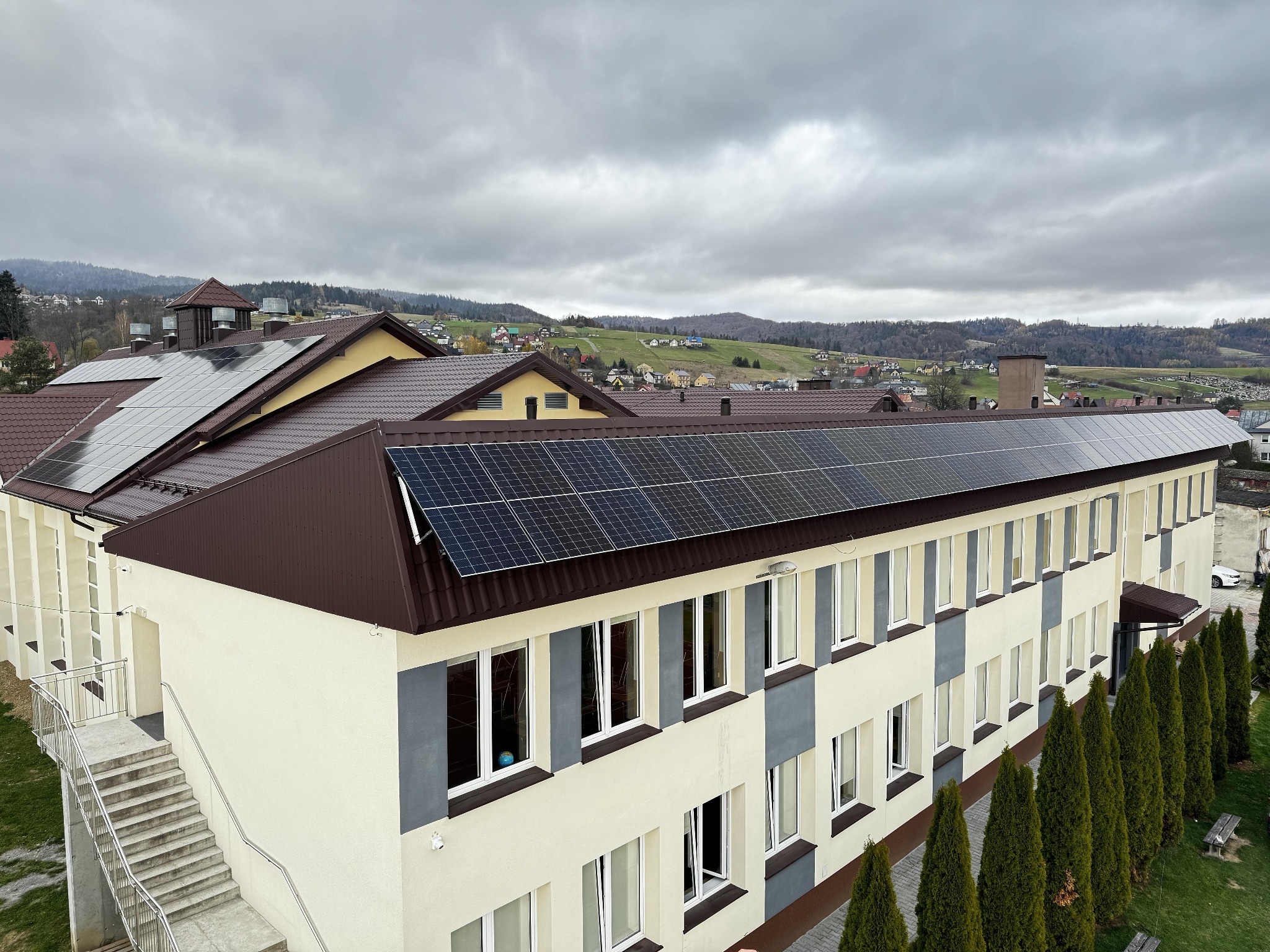 Długi budynek z brązowym dachem i panelami słonecznymi, widok z góry na tle pagórkowatego krajobrazu pod pochmurnym niebem.