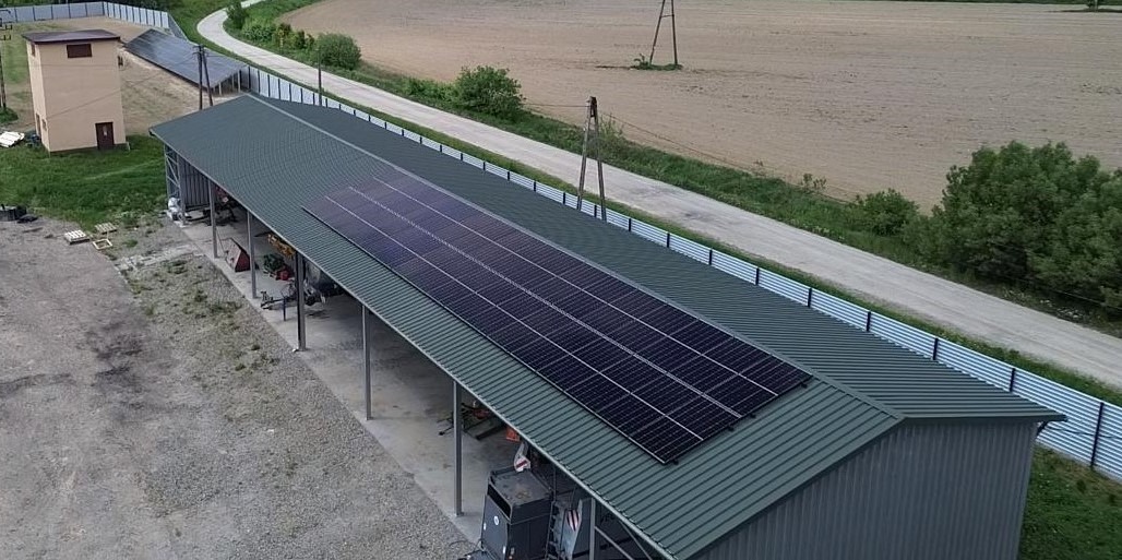 Instalacja paneli na dachu wiaty z blachy trapezowej, widok z góry, obok droga polna i budynek gospodarczy. Gotowa instalacja fotowoltaiczna.