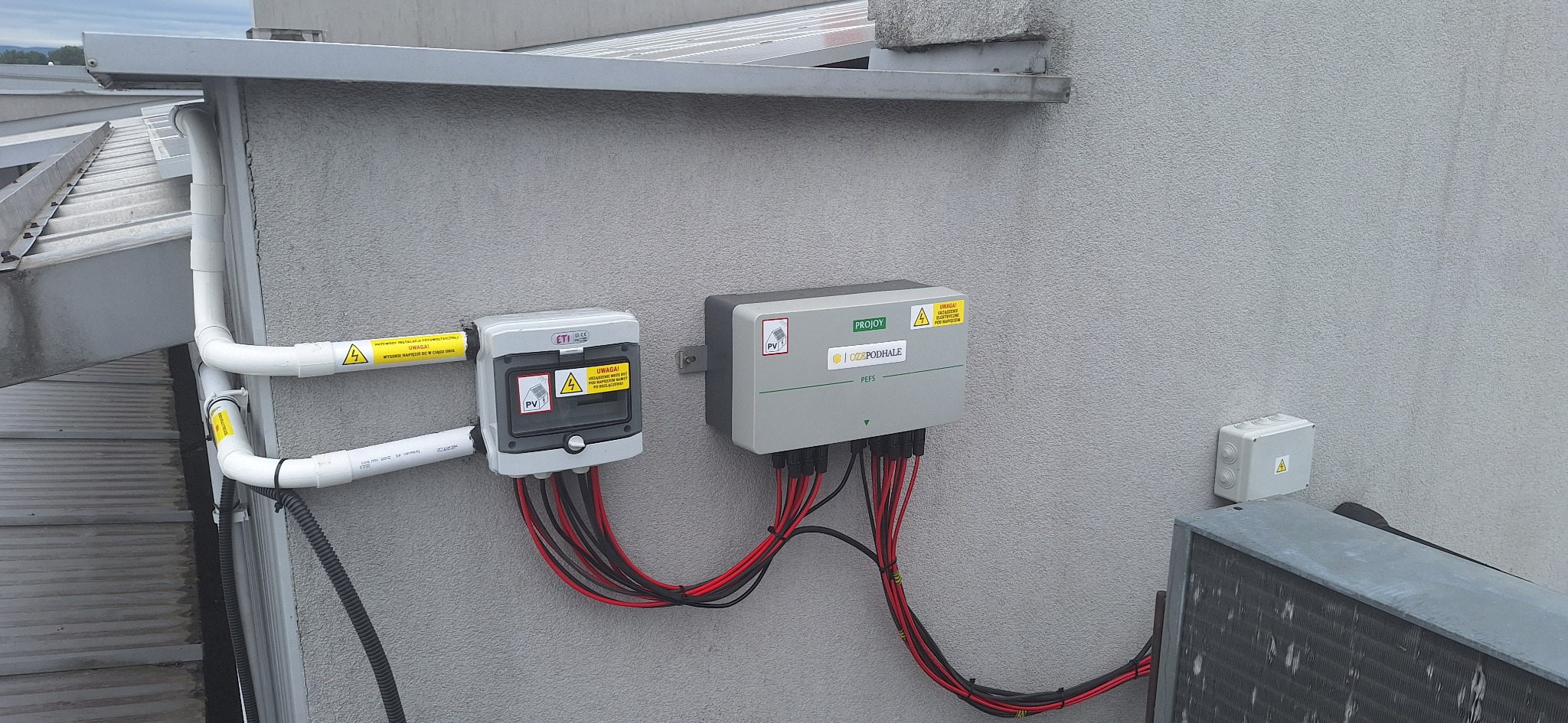 Szara ściana z zamontowanymi elementami instalacji fotowoltaicznej: puszkami ETI, inwerterem PROJOY, rurami PCV i przewodami w izolacji czerwono-czarnej.