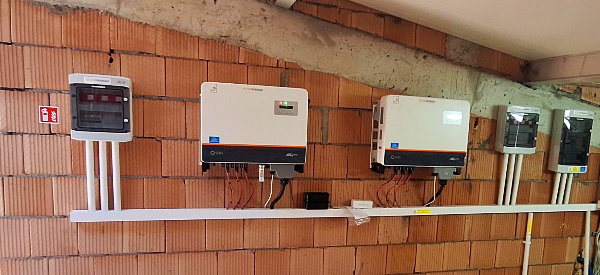 Instalacja elektryczna: dwa inwertery i rozdzielnice na ceglanej ścianie, rury PCV prowadzone wzdłuż ściany. Widoczny znak ostrzegawczy i gaśnica.