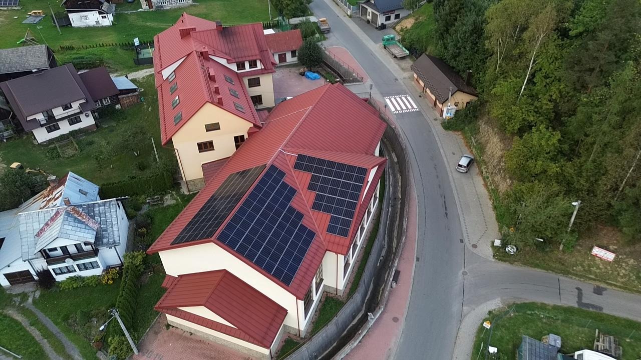Widok z góry na budynek z czerwonym dachem, na którym zamontowane są panele słoneczne. W tle zabudowa wiejska i droga.