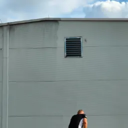 Pracownik w czarno-białej koszulce czyści szarą elewację budynku przemysłowego za pomocą myjki ciśnieniowej, widoczny kontrast między umytą a brudną częścią ściany, w tle niebieskie niebo z białymi...